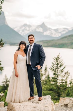 Happy couple stands on a rock with a breathtaking mountain lake vista behind them on their wedding day.