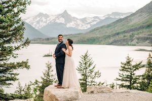 Bride and groom pose on boulder with stunning mountain lake backdrop at outdoor wedding.