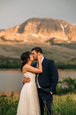 Bride and groom share passionate kiss at sunset with glowing mountain and lake behind them.