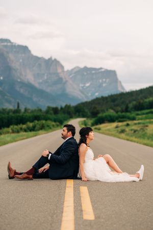 Wedding couple sits back-to-back on a scenic mountain road with stunning rocky peaks in the background.