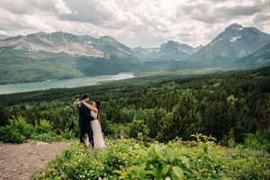 Wedding couple embracing on a hilltop overlooking a stunning mountain lake and forest landscape.