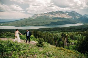 Bride and groom hold hands walking on mountain hillside with scenic lake and peaks behind.