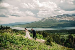 Bride and groom walk hand in hand on a mountain overlook with a lake and peaks behind them.