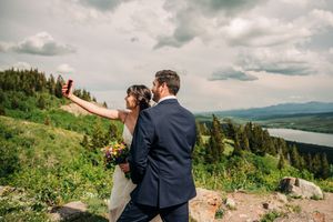 Bride takes selfie with groom on mountain overlook with lush green valley and lake below.