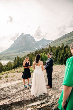 Outdoor wedding ceremony on rocky mountain overlook with officiant, couple, and scenic lake and peak views.