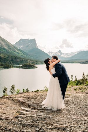 Bride and groom sharing first kiss on rocky overlook with dramatic mountain lake scenery.