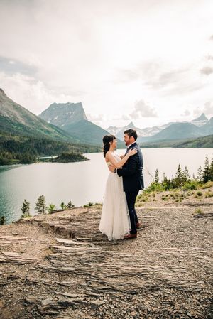 Bride and groom embrace on rocky overlook with breathtaking turquoise mountain lake backdrop.
