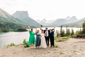 Joyful wedding party celebrates with raised arms against a stunning mountain lake backdrop outdoors.
