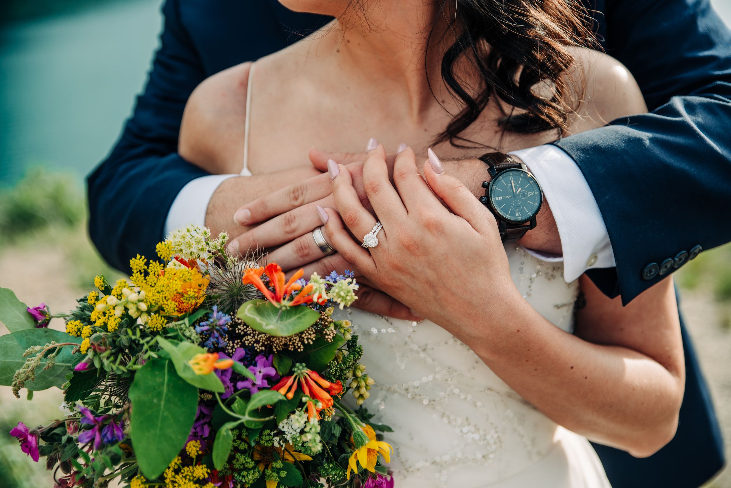 Close-up of groom embracing bride, showcasing wedding rings and vibrant wildflower bouquet.