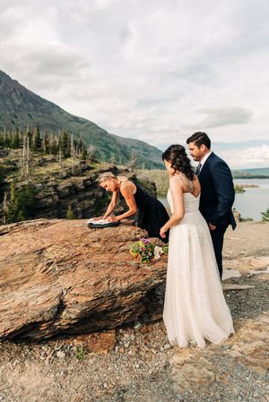 Officiant signs marriage documents on boulder as bride and groom stand at mountain lake wedding.