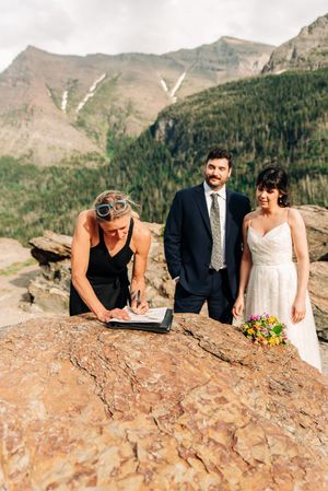 Officiant signs marriage certificate on boulder as smiling couple watches in mountains.