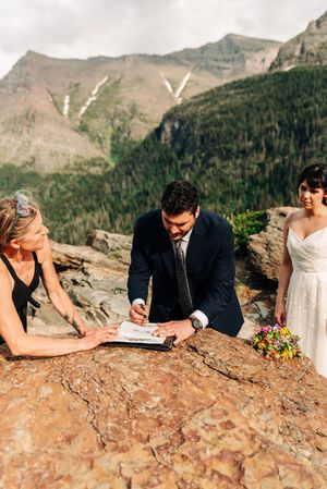 Groom signs wedding license on a rocky surface with mountains behind as bride and officiant watch.