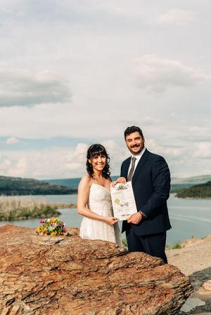 Smiling newlyweds display marriage certificate on rocky overlook with lake and sky behind them.