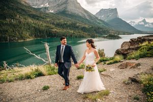 Bride and groom walk hand in hand along a scenic mountain trail beside a vivid turquoise lake.