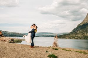 Groom lifts bride in a joyful embrace on a rocky shoreline with a scenic mountain lake behind them.