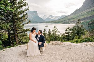 Bride and groom laughing together on rocky overlook with stunning mountain lake panorama.