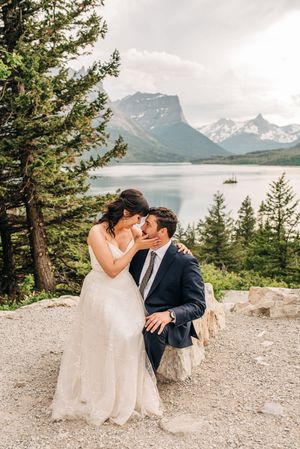 Bride leaning in to kiss groom seated on rock with pine trees and mountain lake behind.
