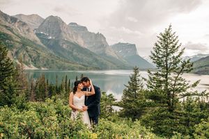Bride and groom embracing among lush greenery with scenic mountain lake and peaks backdrop.