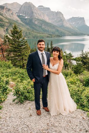 Bride and groom pose together among lush greenery with a serene mountain lake and peaks behind them.