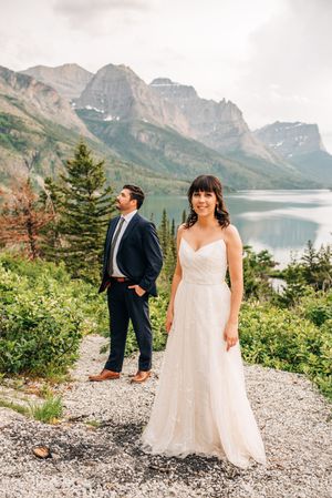 Bride smiles in foreground while groom stands behind her with a stunning mountain lake backdrop.