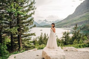 Bride in white gown stands on rocky ledge with majestic mountain lake and peaks behind her.