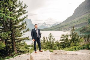Groom in navy suit stands on rocky overlook with stunning mountain lake views at Glacier National Park.