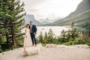 Bride and groom share romantic moment on rocky overlook above turquoise lake in Glacier National Park.