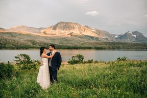Bride and groom embrace in lush green meadow with stunning mountain lake backdrop during golden hour outdoor wedding.