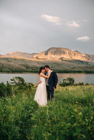 Bride and groom embrace tenderly in a lush green meadow with a glowing mountain and lake at sunset.