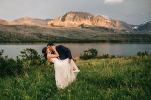 Couple sharing a romantic dip kiss in lush green meadow with mountain lake backdrop during golden hour wedding photography.