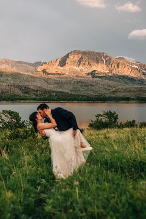 Groom lifts bride for a romantic kiss in a green meadow with a dramatic mountain lake at golden hour.