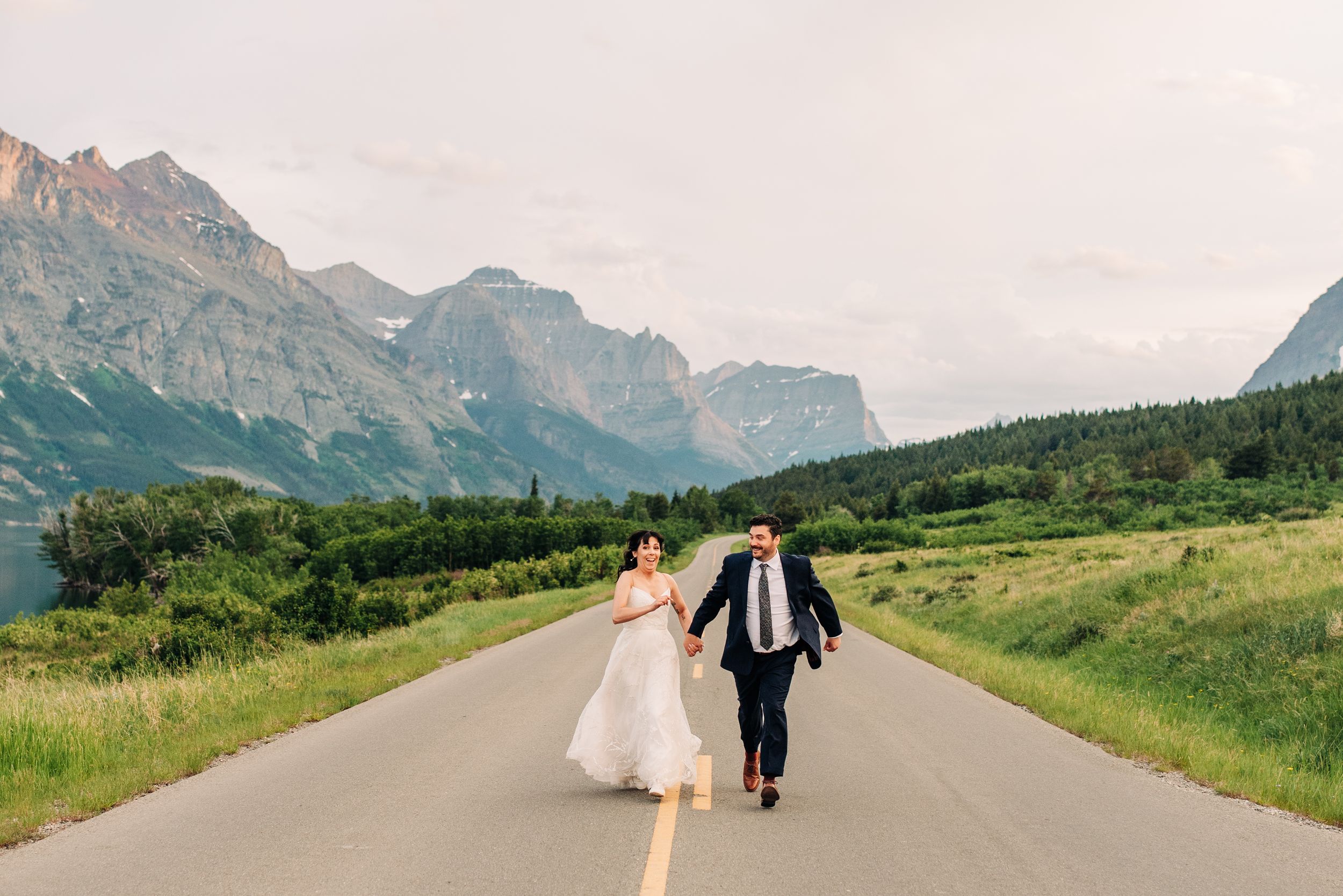 Bride and groom walking hand-in-hand down an open road surrounded by majestic mountain scenery.