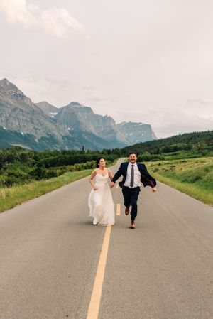 Laughing newlyweds run hand in hand down an open road framed by mountains and greenery.
