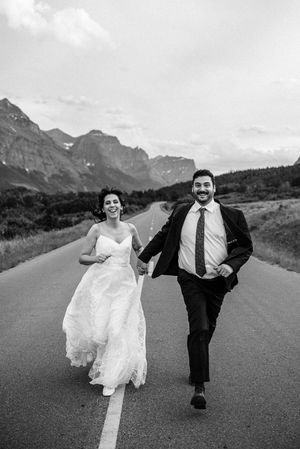 Laughing bride and groom run down an open road with mountains in the background in black and white.