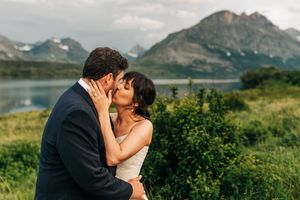 Bride and groom share a tender kiss beside a mountain lake surrounded by lush green foliage.