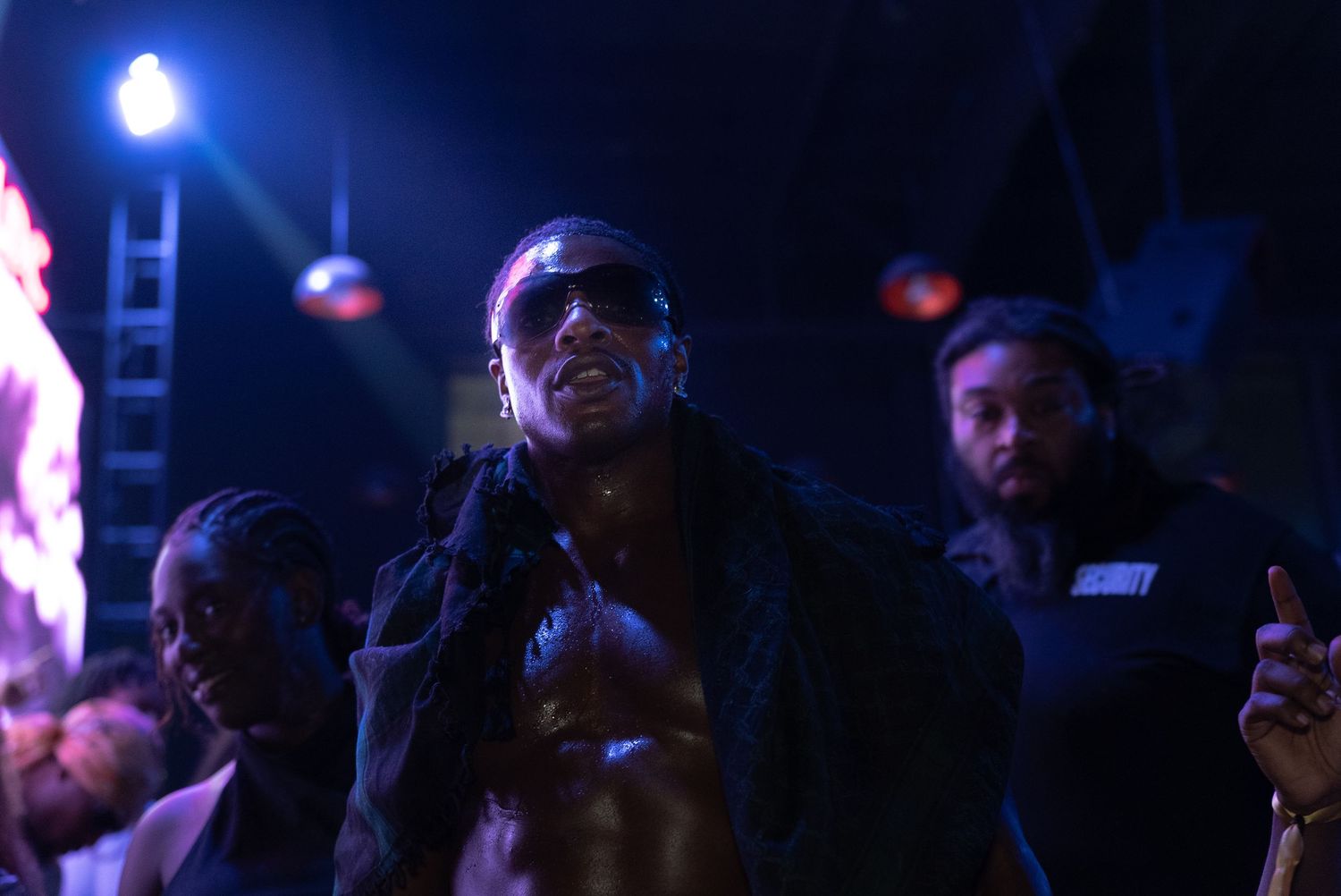 Man wearing sunglasses performing at a nightclub event with crowd and purple stage lighting.