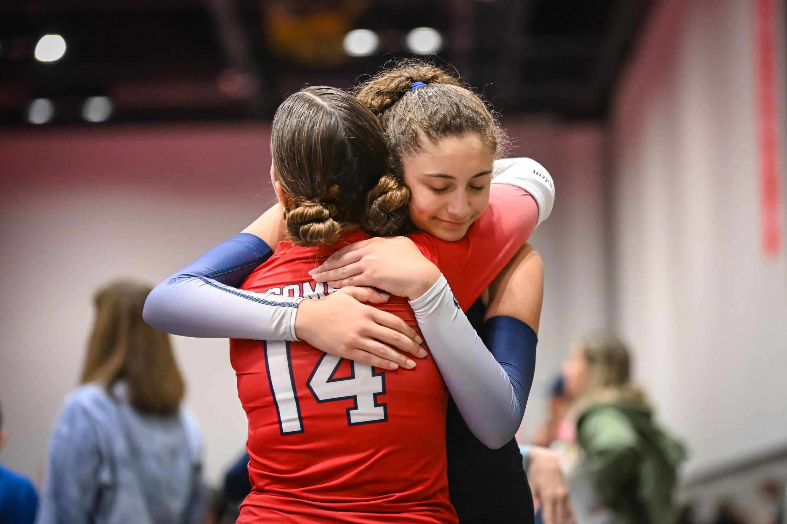 Two volleyball players in red uniforms share a warm embrace during a tournament game.