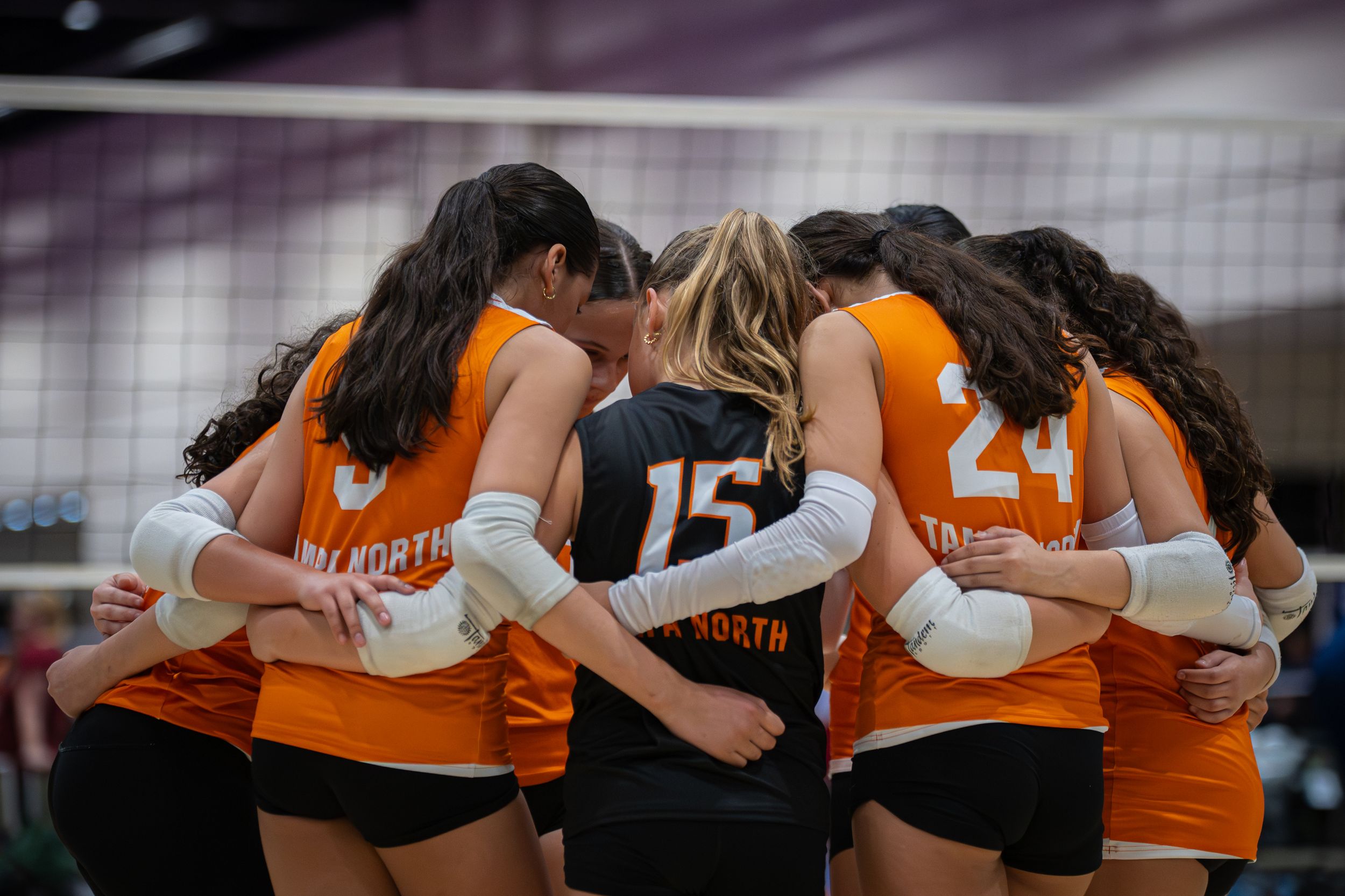 Girls volleyball team in orange jerseys huddle together on court during a competitive match.