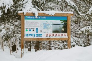 Hardy Lake Trails informational sign board surrounded by snow-covered trees in winter.