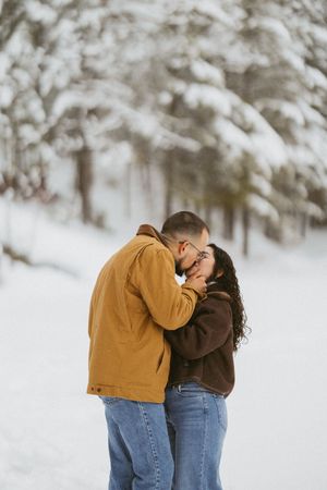 Romantic couple sharing a kiss in snowy forest setting, wearing casual winter jackets.