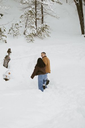 Couple walking through deep snow in a winter forest, wearing brown and blue jeans outfits.