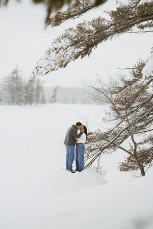 Couple sharing a romantic kiss in a snowy winter landscape surrounded by snow-covered pine trees.