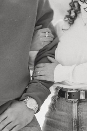 Black and white photo of couple holding hands, showing engagement ring and watch, wearing jeans and casual sweaters.