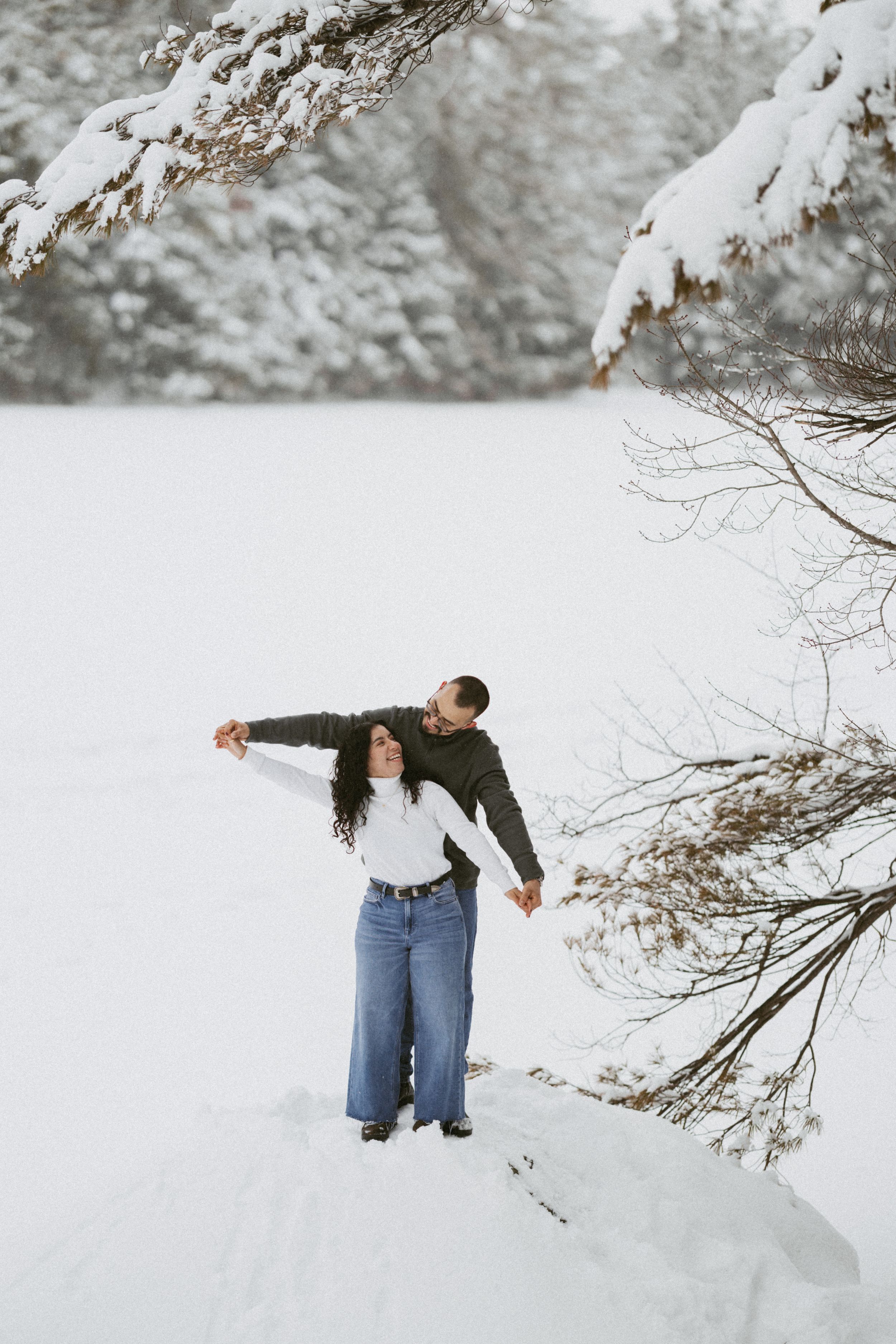 Romantic couple embracing in a snowy winter landscape, woman in white top and wide-leg jeans.