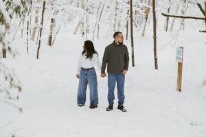 Couple holding hands on a snowy forest path in winter, wearing jeans and casual sweaters surrounded by snow-covered trees.