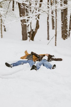 Couple making snow angels together in a snowy forest, wearing winter jackets and jeans.