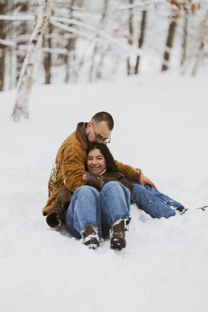 Happy couple sitting together in deep snow in a winter forest, man hugging woman from behind.