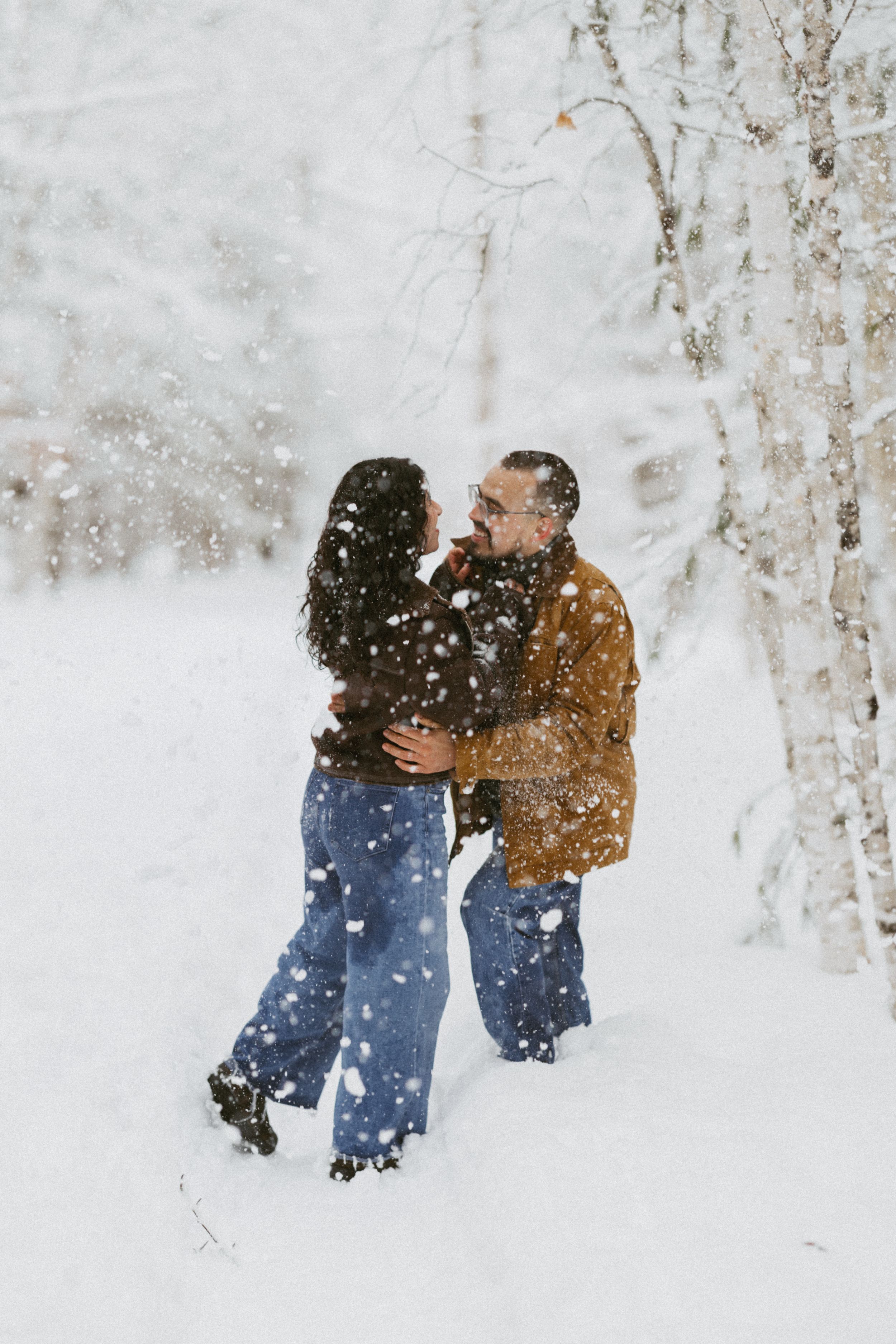 Couple dancing and laughing in falling snow surrounded by snow-covered trees in winter.