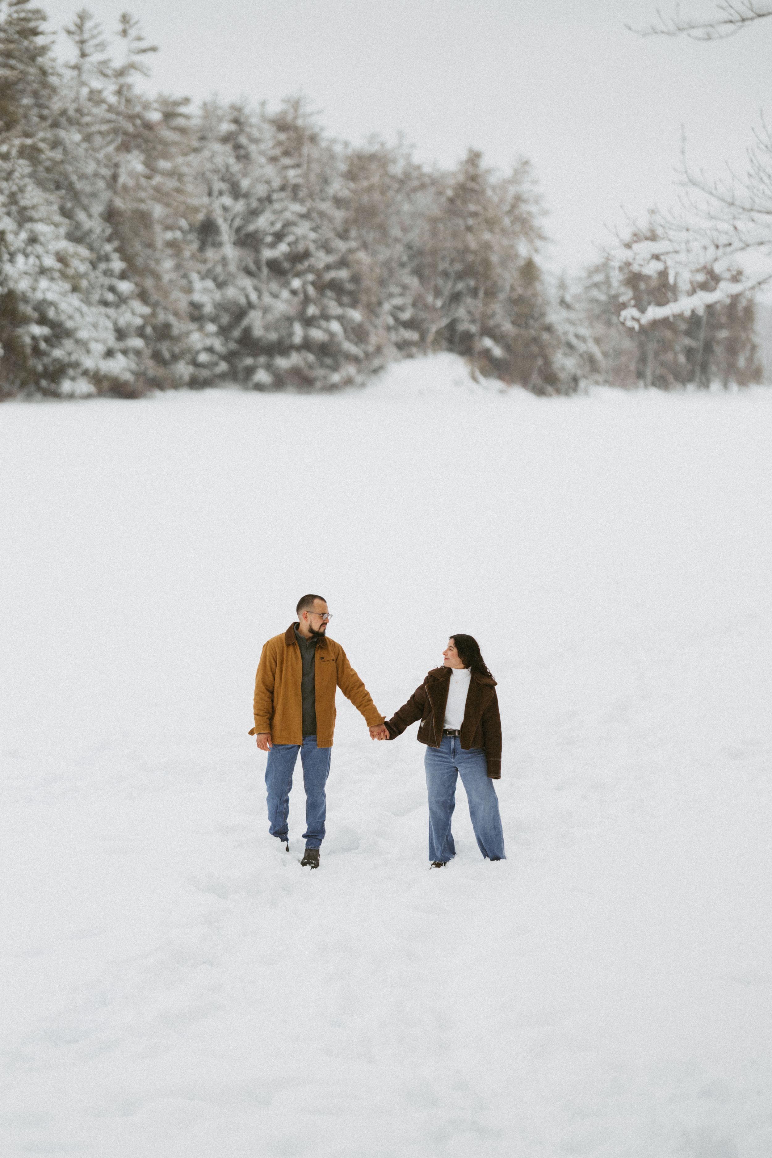 Couple holding hands walking through snowy winter landscape with snow-covered trees in background.