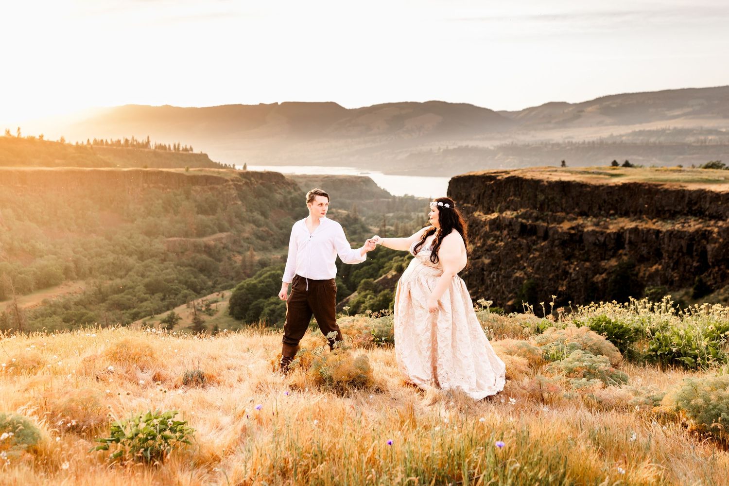 Couple holds hands at golden hour on a scenic cliff overlooking a river valley with mountains in the background.